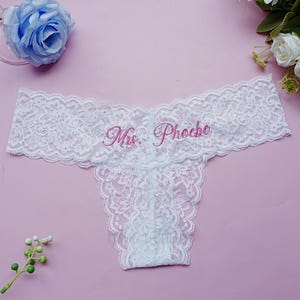 May include: White lace thong underwear with the embroidered text "Mrs. Phoebe" in pink. The delicate lingerie features a floral lace pattern and a low-rise design, suitable for bridal wear or a special occasion.