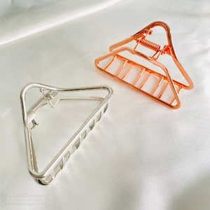 Puede incluir: Dos pinzas para el pelo de metal. Una es plateada, la otra es oro rosa. Ambas son triangulares con una bisagra de resorte. Las pinzas están sobre una superficie blanca y sedosa.