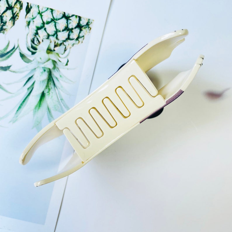 May include: A white plastic hair claw with a row of teeth on the underside. The claw is open and ready to hold hair.