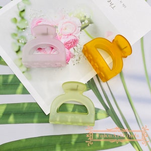 Puede incluir: Tres pinzas para el pelo de plástico en diferentes colores: rosa claro, verde claro y amarillo. Las pinzas tienen forma de semicírculo con la parte superior redondeada y la inferior plana.