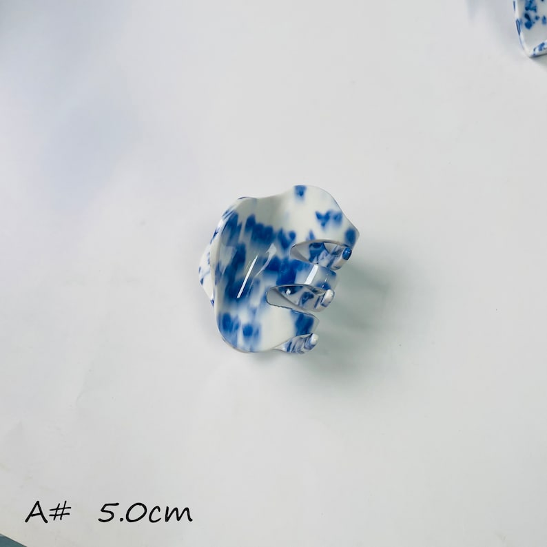 K&ouml;nnte beinhalten: Eine wei&szlig;e und blaue Haarklammer mit einem blauen und wei&szlig;en Wirbelmuster. Die Klammer ist 5,0 cm lang.