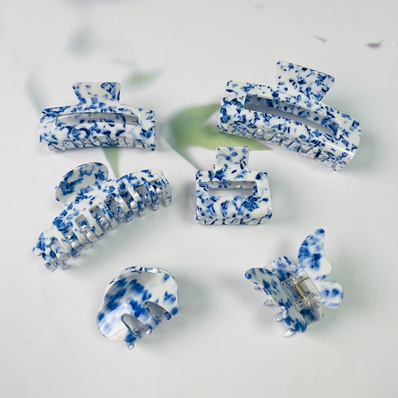 K&ouml;nnte beinhalten: Satz mit sechs blauen und wei&szlig;en gemusterten Haarklammern. Die Klammern sind aus Kunststoff und haben verschiedene Formen, darunter Schmetterling, Rechteck und Klammer.