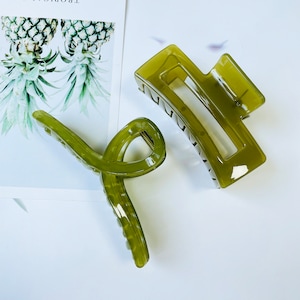 Puede incluir: Dos pinzas para el pelo de plástico verde. Una pinza es un rectángulo grande con la parte superior curvada y un rectángulo más pequeño cortado en el medio. La otra pinza es una forma larga y curva con un pequeño tornillo de metal en el centro.