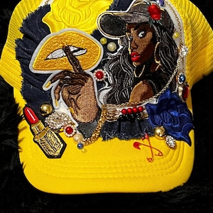 May include: Yellow trucker hat with a mesh back and a decorative front. The front features an embroidered image of a woman, a gold lip design, a lipstick, and various embellishments including pearls and jewels.