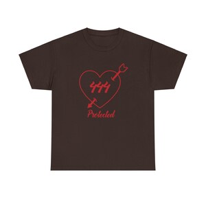 Könnte beinhalten: Braunes T-Shirt mit einem roten Herz, das von einem Pfeil durchbohrt wird. Das Herz enthält die Zahl "444" und das Wort "Protected" darunter. Das Design ist auf der Vorderseite des T-Shirts aufgedruckt.