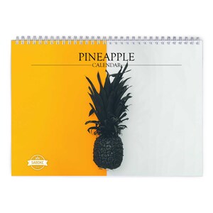 Peut inclure: Un calendrier mural à reliure spirale avec un fond bicolore, jaune vif à gauche et blanc à droite. Un graphique d'ananas noir est centré au milieu. Le texte PINEAPPLE CALENDAR est imprimé en noir, accompagné d'un logo SABOKE.