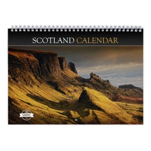 Könnte beinhalten: Ein spiralgebundener Wandkalender mit dem Titel SCOTLAND CALENDAR in weißer und goldener Schrift. Das Deckblatt zeigt eine Landschaftsaufnahme von hügeligem Grasland und Felsklippen unter einem bewölkten Himmel sowie ein kleines SABOKE-Logo.