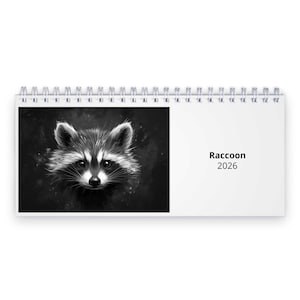 Puede incluir: Calendario de escritorio con espiral que presenta una ilustración en blanco y negro de un mapache. La cara del mapache está en la página izquierda, con la palabra "Raccoon" y el año "2026" impresos en la página derecha.