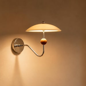Op de afbeelding: Een wandlamp met een gebogen chromen arm, een klein houten bolvormig accent en een brede, ondiepe, gebroken witte koepelvormige kap die een warm, zacht licht uitstraalt, bevestigd aan een ronde chromen basisplaat op een neutrale muur.