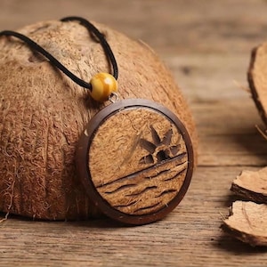 May include: A round, wooden pendant necklace with a carved tropical scene of a palm tree and waves. The pendant hangs from a black cord with a yellow bead. The necklace is displayed on a coconut shell, with pieces of the shell nearby.