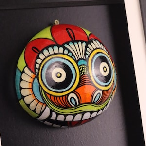 May include: A hand-painted decorative mask with a quirky design. The mask is round with large eyes, a wide grin, and bright colours including red, yellow, green, and orange. It is mounted on a black background. The mask is likely made of wood.