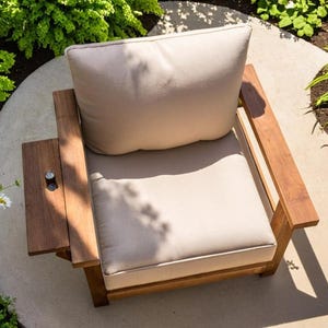 K&ouml;nnte beinhalten: Ein Holz-Au&szlig;enstuhl mit hellbeige Kissen und R&uuml;ckenlehne. Der Stuhl hat einen eingebauten Beistelltisch. Der Stuhl ist von Gr&uuml;n und Blumen umgeben. Der Stuhl hat ein modernes Design.