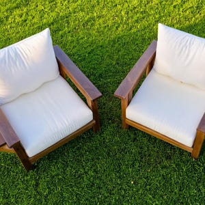 K&ouml;nnte beinhalten: Zwei Holz-Au&szlig;ensessel mit wei&szlig;en Kissen und Armlehnen. Jeder Stuhl hat einen Beistelltisch. Die St&uuml;hle stehen auf einem gr&uuml;nen Rasen und schaffen einen entspannten Sitzbereich im Freien.