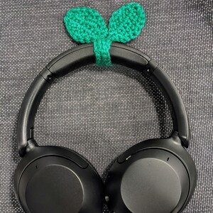 May include: Black over-ear headphones with a green, crocheted leaf design attached to the headband. The headphones have large, round earcups and a padded headband. The crocheted leaves are a bright green colour.