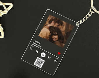 Puede incluir: Un llavero de acrílico transparente con una imagen en blanco y negro de una pareja. El llavero tiene un diseño de reproductor de música con el título de la canción "Daylight" de David Kushner, un botón de reproducción y un código QR.