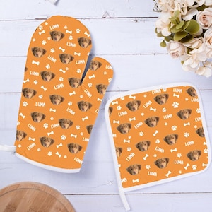 Op de afbeelding: Een set van twee oranje ovenwantjes met een witte rand. De wantjes hebben een herhalend patroon van een hondenkop en de naam "Luma" met witte poot afdrukken en botvormen.