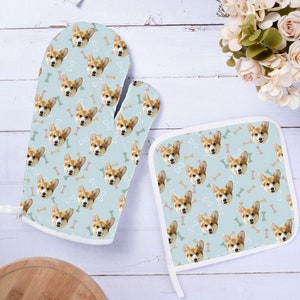 Könnte beinhalten: Ein Set aus zwei blauen Ofenhandschuhen mit weißem Rand. Die Handschuhe haben ein sich wiederholendes Muster aus einem Hundegesicht und Knochenformen. Das Hundegesicht ist hellbraun mit weißem Fell.