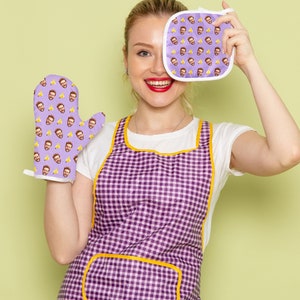 Op de afbeelding: Een vrouw draagt een paars-gele geruite schort en houdt een paarse ovenwant vast met een herhaald patroon van een mannengezicht. De ovenwant bedekt haar rechteroog. Ze draagt ook een wit T-shirt.