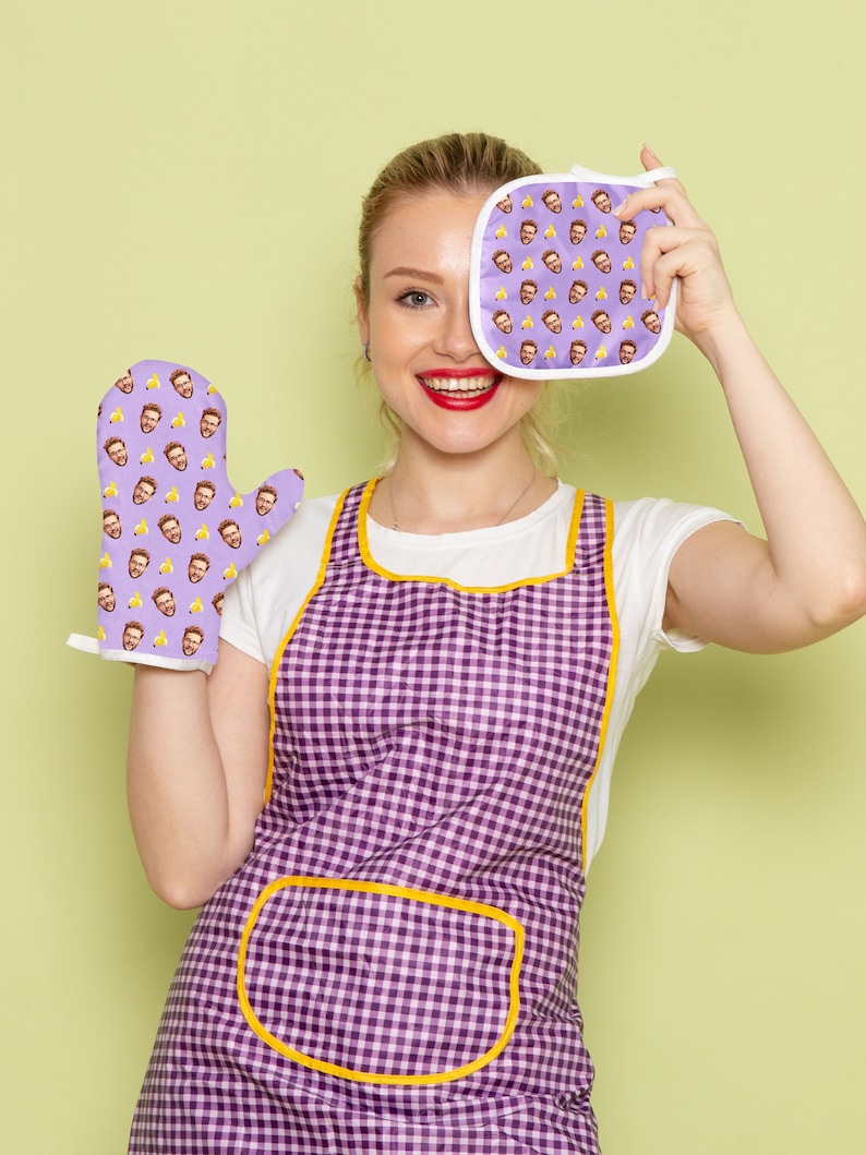 Op de afbeelding: Een vrouw draagt een paars-gele geruite schort en houdt een paarse ovenwant vast met een herhaald patroon van een mannengezicht. De ovenwant bedekt haar rechteroog. Ze draagt ook een wit T-shirt.