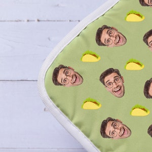 Op de afbeelding: Een groene laptophuls van stof met een witte rits en een herhalend patroon van een mensen gezicht en een gele taco.