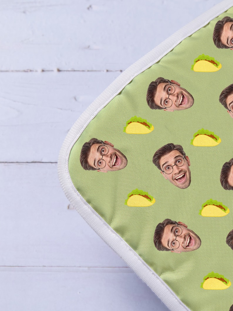 Op de afbeelding: Een groene laptophuls van stof met een witte rits en een herhalend patroon van een mensen gezicht en een gele taco.