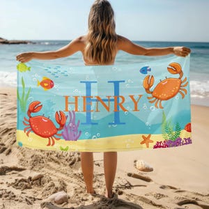Op de afbeelding: Een gepersonaliseerde strandlaken met een kleurrijke onderwaterwereld, inclusief cartoonkrabben, vissen en zeewier. De naam "HENRY" staat in oranje letters op een blauwe achtergrond, perfect voor een dagje strand.