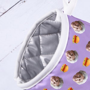 Op de afbeelding: Paarse ovenwant met een witte gewatteerde voering en een witte ophanglus. De want is bedekt met een herhalend patroon van een mensen gezicht en een hamburger.