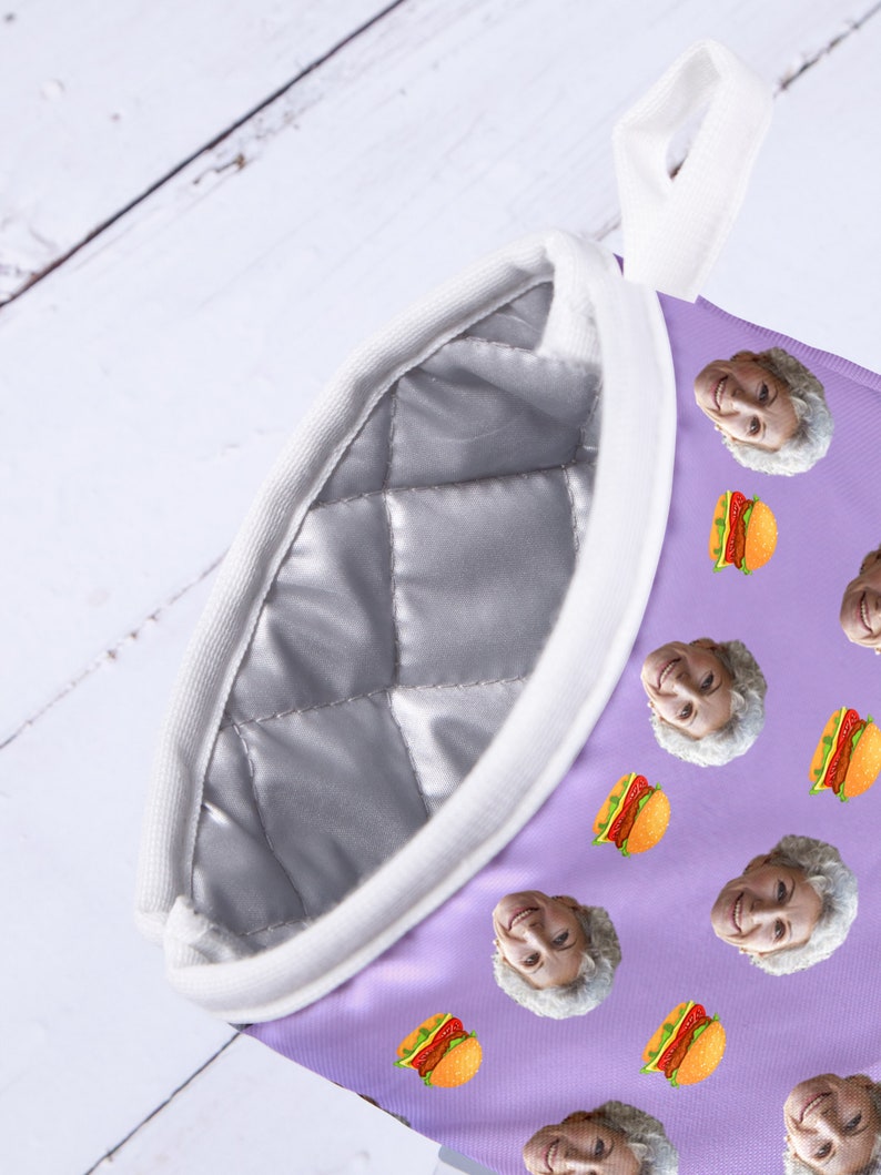 Op de afbeelding: Paarse ovenwant met een witte gewatteerde voering en een witte ophanglus. De want is bedekt met een herhalend patroon van een mensen gezicht en een hamburger.