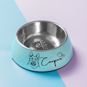 May include: A light blue dog bowl with a stainless steel interior. The bowl has a black silhouette of a German Shepherd dog and the name "Cooper" printed on the side.