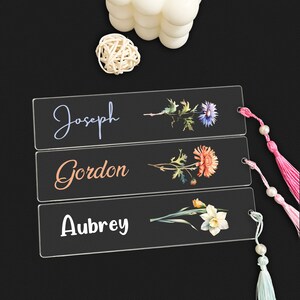 May include: Three clear acrylic bookmarks with floral designs and personalised names. The top bookmark has a blue flower and the name "Joseph". The middle bookmark has an orange flower and the name "Gordon". The bottom bookmark has a white flower and the name "Aubrey".