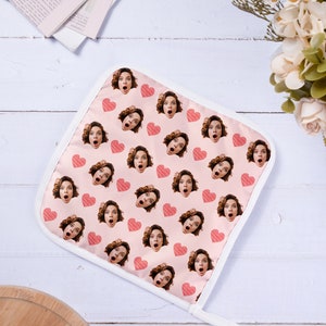 Op de afbeelding: Een roze ovenwant met een witte rand en een herhalend patroon van een vrouwen gezicht met een verraste uitdrukking en een rood hart.