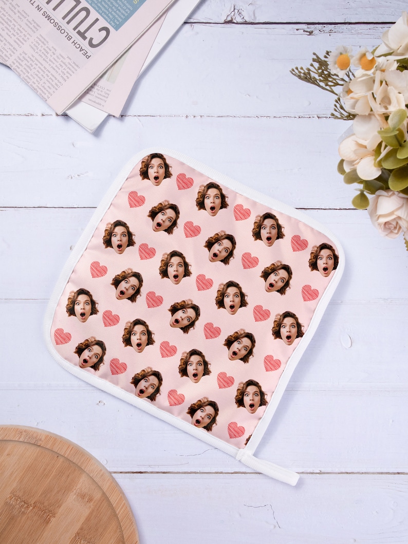 Op de afbeelding: Een roze ovenwant met een witte rand en een herhalend patroon van een vrouwen gezicht met een verraste uitdrukking en een rood hart.