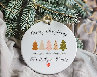 May include: A white ceramic Christmas ornament with the text "Merry Christmas" and "The Wilson Family." It features five illustrated Christmas trees in various colors and a small red heart. A jute string is attached for hanging.