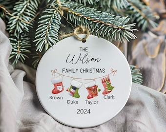 May include: White ceramic Christmas ornament with the text "The Wilson Family Christmas 2024" and four stockings with names "Brown", "Duke", "Taylor", and "Clark" hanging from a string.