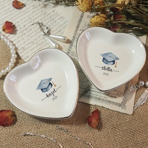 May include: Two heart-shaped, white ceramic dishes with gold trim. Each dish features a watercolor graduation cap design and a name with the year 2026. One dish says "Hazel" and the other says "Stella".