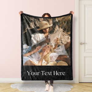 May include: A black fleece blanket with a photo of two people sitting on a blanket in a field. The photo shows a person wearing a straw hat reading a book to another person. The text "Your Text Here" is printed on the blanket.
