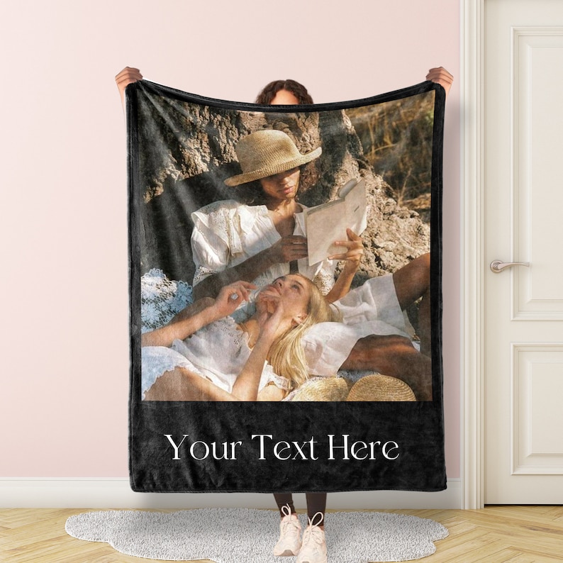 May include: A black fleece blanket with a photo of two people sitting on a blanket in a field. The photo shows a person wearing a straw hat reading a book to another person. The text "Your Text Here" is printed on the blanket.