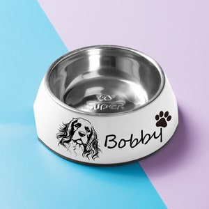 May include: White dog bowl with a stainless steel interior. The bowl has a black paw print and the name "Bobby" printed on the side. A black and white illustration of a dog's head is also printed on the bowl.