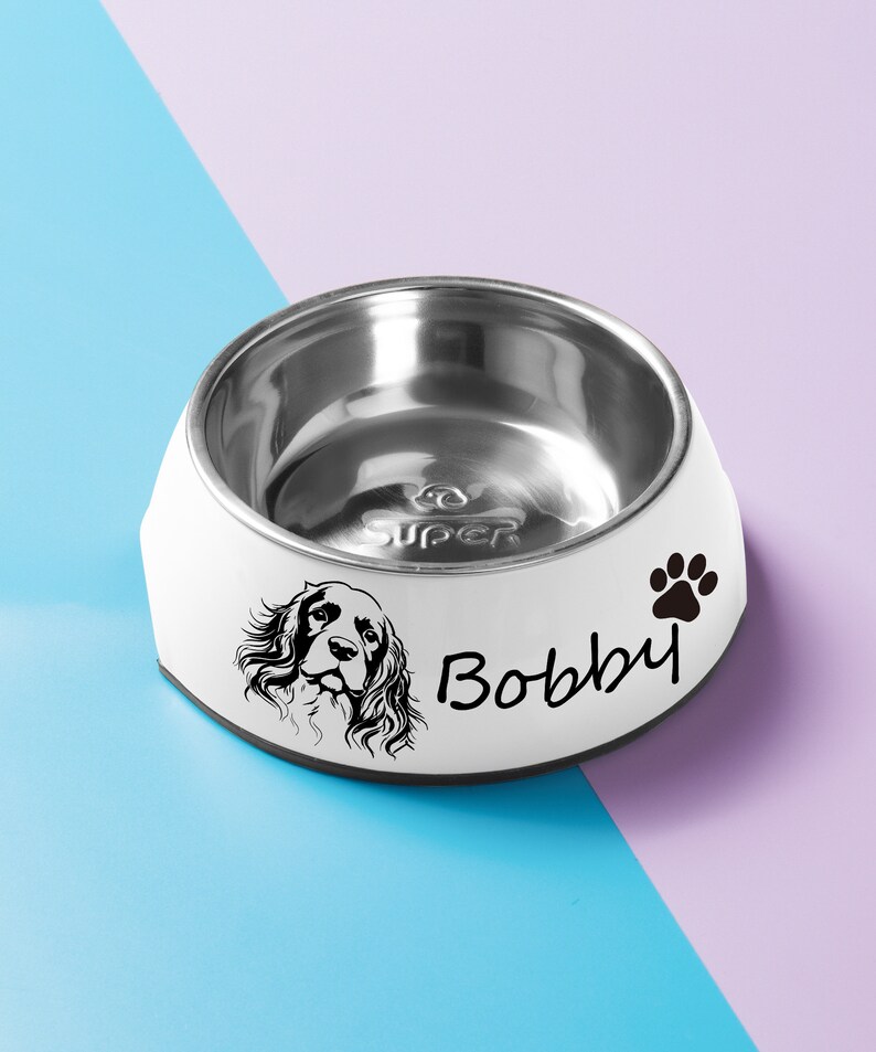 May include: White dog bowl with a stainless steel interior. The bowl has a black paw print and the name "Bobby" printed on the side. A black and white illustration of a dog's head is also printed on the bowl.