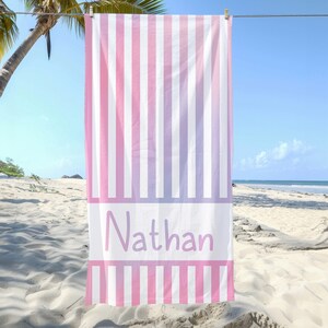 Puede incluir: Una toalla de playa a rayas rosa y blanca con el nombre "Nathan" impreso en morado en la parte delantera.