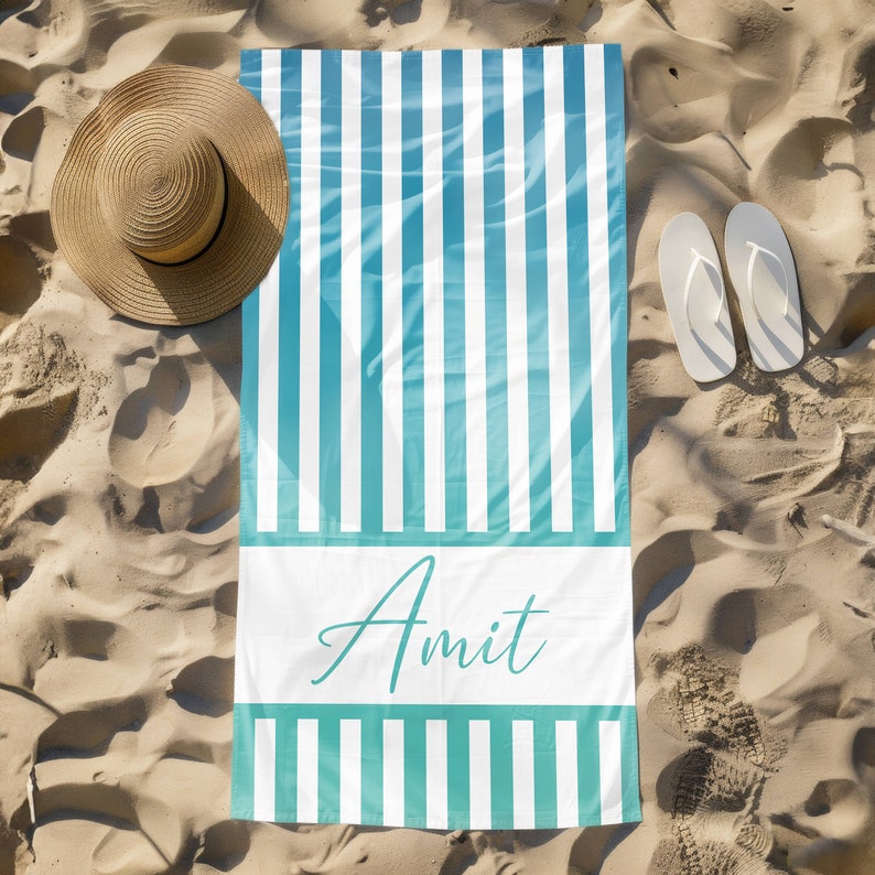 Puede incluir: Una toalla de playa blanca con rayas azules y blancas y el nombre "Amit" impreso en letra cursiva. La toalla est&aacute; sobre una playa de arena.