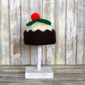 May include: A knitted Christmas pudding hat with a red pom-pom and green holly leaves. The hat is displayed on a white stand.