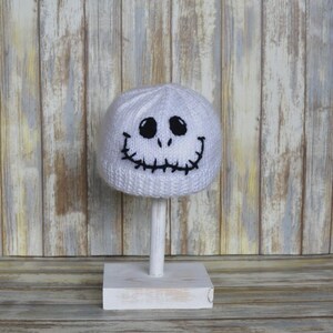 May include: White knitted hat with a black embroidered Jack Skellington face. The hat is displayed on a white stand.