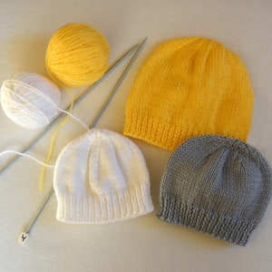 May include: Three knitted hats in different colors: yellow, white, and gray. The hats are lying on a white surface with two knitting needles and balls of yarn.