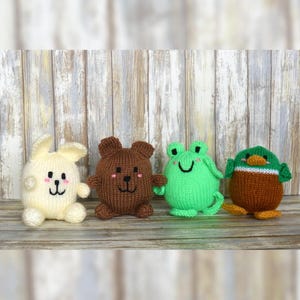 May include: Four knitted toy animals: a cream-coloured rabbit, a brown bear, a green frog, and a brown and green duck. Each has embroidered features and sits on a wooden surface. The rabbit has long ears, and the duck has a green head.