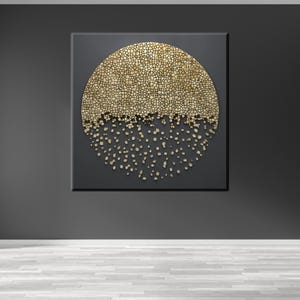 以下が含まれることがあります： 金色と黒色の抽象的な壁掛けアート作品で、円形のデザインと質感のある表面が特徴です。円の上が無垢の金で、下がより小さな金色の破片でできており、落下しているように見えます。