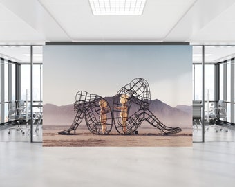 Op de afbeelding: Een zwart metalen sculptuur van twee figuren, een groter dan de andere, zittend in een woestijnlandschap. De figuren zijn gemaakt van draadgaas en lijken elkaar te omhelzen.