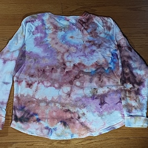 May include: A long-sleeved shirt with a tie-dye pattern in shades of pink, purple, blue, and brown. The shirt has a relaxed fit and a crew neck.