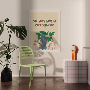 May include: A framed print with the text "You don't have to earn self-care" in colorful letters. The print features a stylized illustration of a plant with a blue figure in the center. The print is hanging on a light beige wall.