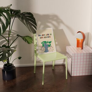 May include: A green chair with a framed print that reads "You don't have to earn self-care" with a cartoon illustration of a plant. The chair is in front of a white wall and a small white and pink tiled table with a lamp on top.
